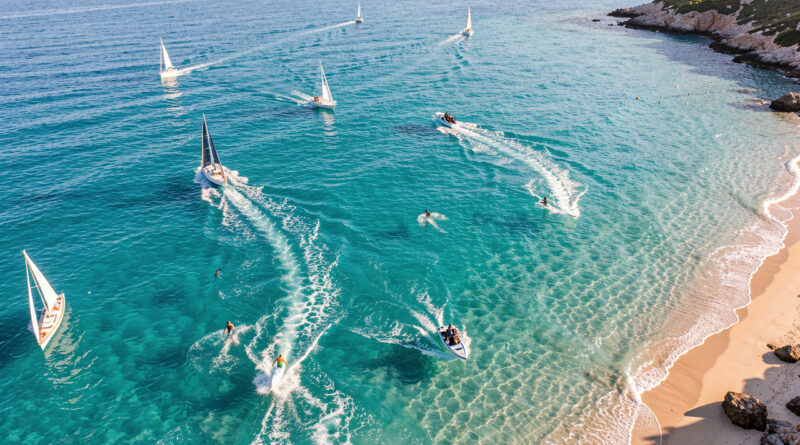 Vue aérienne en été en Corse sur une eau turquoise, montrant plusieurs activités nautiques sportives comme des bateaux à voile et des traces de vitesse.
